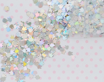 May include: A collection of small, iridescent, holographic bunny-shaped sequins scattered across a surface. The individual bunny shapes feature a reflective, multi-colored finish that shifts in light, creating a shimmering effect across the pile.