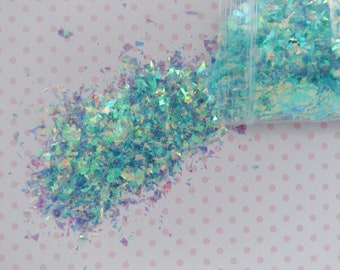 May include: A pile of iridescent light blue and teal craft flakes spilling from a clear plastic bag. The thin, jagged, reflective pieces exhibit a color-shifting effect, displaying hints of purple and green when catching the light.