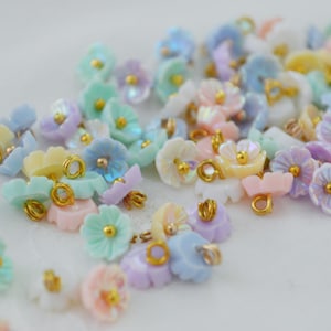 May include: A collection of small, multi-colored flower-shaped charms in pastel shades of pink, blue, yellow, and green. Each charm features a central gold-toned bead and a small gold-toned metal loop attached to the back for hanging or connecting.