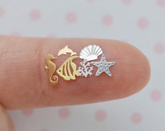 May include: A collection of miniature metal charms featuring various sea life shapes including a gold seahorse, a gold fish, a small gold dolphin, a silver seashell, a silver coral branch, and a silver starfish, all arranged together in a small cluster.