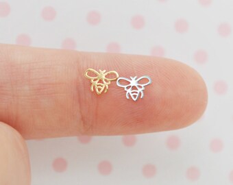 May include: Two small bee-shaped charms, one in a gold tone and one in a silver tone. Each charm features a cut-out design representing the wings and body of a bee. These tiny metal pieces are flat and display a detailed outline of the insect.