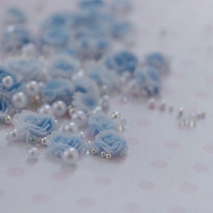 May include: A collection of light blue fabric roses, white pearls, and small silver beads scattered on a pink polka dot surface. The roses are clustered together, with some pearls and beads interspersed among them.