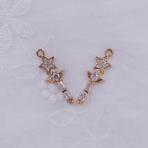 May include: Two gold-colored charms in the shape of a star and a wand. The star is encrusted with small, clear crystals. The wand has a clear crystal and rectangular crystals. The charms have a small loop at the top for attaching to jewelry.