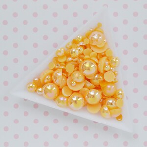 May include: A white triangular tray filled with a collection of small, round, orange beads. The beads feature a mix of matte and iridescent finishes, with varying sizes of spherical and half-spherical shapes clustered together in the container.