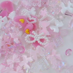 May include: A collection of pink and white decorative embellishments, including hearts, bows, and beads. Some pieces have iridescent finishes, and others feature yellow accents. The assortment is scattered across a light background.