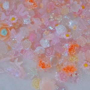 May include: A collection of small, decorative resin cabochons in various shapes including roses, flowers, bows, and starfish. The pieces feature a palette of translucent pink, peach, orange, and iridescent white tones with glossy finishes.