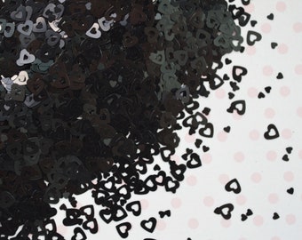 May include: A large pile of small, black, heart-shaped confetti pieces. Each individual piece is a hollow outline of a heart. The confetti is scattered across the surface, with a dense cluster on the left side and individual hearts spread out.