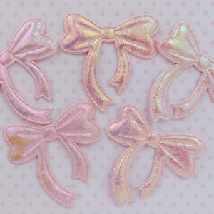 May include: Five individual bow-shaped patches featuring a shimmering, iridescent pink finish. Each patch has a textured surface with a metallic sheen and stitched edges, arranged in a scattered pattern across the frame for clear visibility.