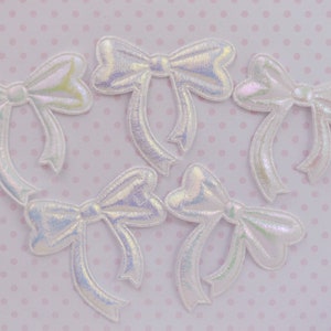 May include: Five iridescent white fabric bows with a 3D effect. The bows are arranged in a cluster on a pink background with white polka dots.