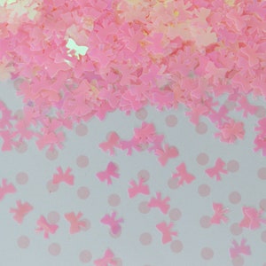 May include: A collection of small, iridescent pink bow-shaped sequins scattered across a surface. The individual bow pieces feature a translucent, light-reflecting finish that creates a soft glow, with some pieces clustered together and others spread out.