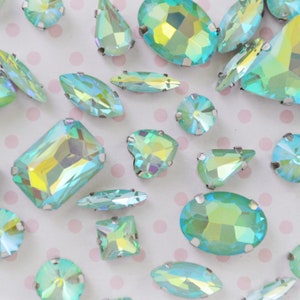 May include: A collection of loose, faceted glass rhinestones in various geometric shapes including ovals, rectangles, triangles, and teardrops. Each stone features an iridescent, light-reflecting finish with prominent teal and yellow hues.
