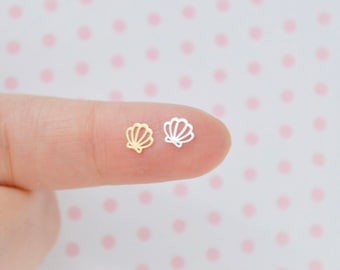 May include: Two small, flat, cutout-style seashell charms, one in a gold tone and one in a silver tone. Each charm features a simple, open-line design depicting the classic fan shape of a scallop shell with vertical ridges radiating from the base.