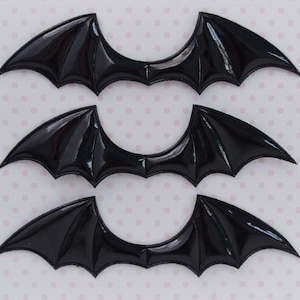 May include: Three identical black bat wing appliques with a glossy, patent-like finish. Each wing features a distinct, pointed shape with raised, molded details that define the structure of the wings, arranged in a vertical row on a light surface.