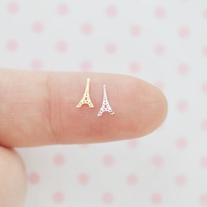 May include: Two miniature Eiffel Tower charms, one in a gold-tone finish and one in a silver-tone finish. Each charm features a delicate, openwork lattice design that mimics the structure of the tower, presented as small metal embellishments.