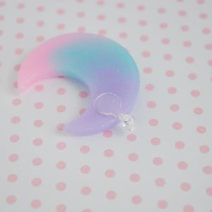 May include: A crescent moon-shaped charm featuring a smooth, glittery gradient finish that transitions from pink to blue to purple. A small, clear plastic jump ring is attached to the lower curve of the moon, providing a functional loop for hanging.