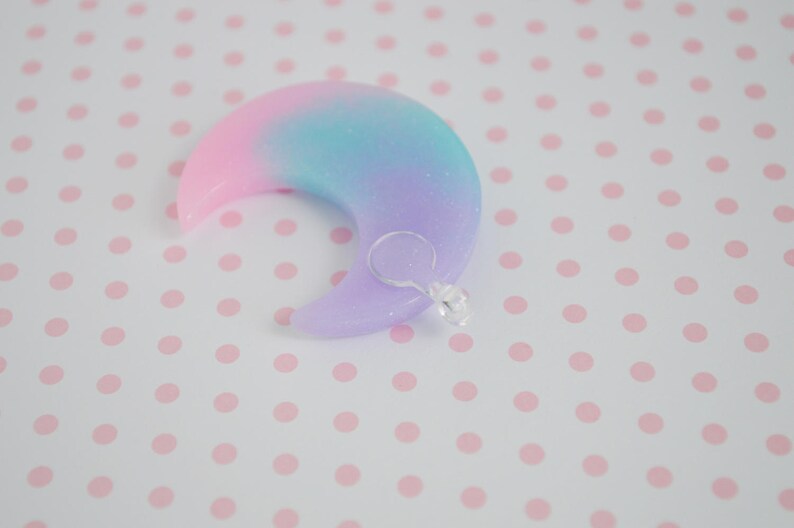 May include: A crescent moon-shaped charm featuring a smooth, glittery gradient finish that transitions from pink to blue to purple. A small, clear plastic jump ring is attached to the lower curve of the moon, providing a functional loop for hanging.