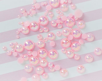 May include: A collection of round, pink, iridescent cabochons in various sizes scattered across a surface. The cabochons feature a smooth, glossy finish with a reflective, pearlescent sheen that shifts in colour, interspersed with smaller matte pink circles.