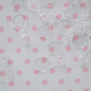 May include: A collection of small, clear plastic circular connectors with a tabbed end. These translucent components feature a round loop attached to a short, solid stem with a small protrusion at the tip, designed for linking or hanging items.