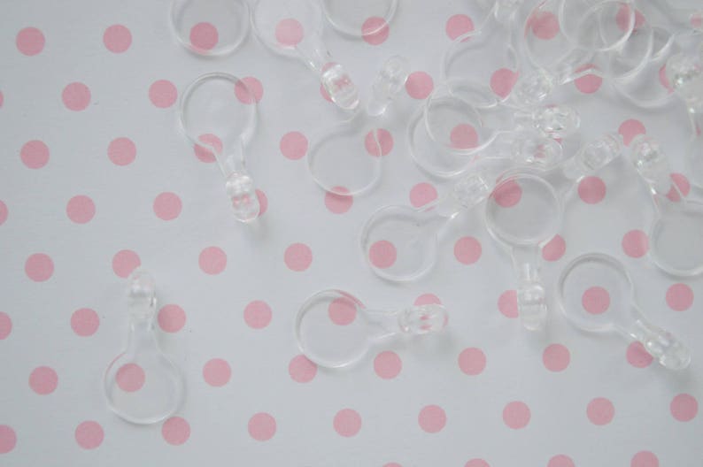 May include: A collection of small, clear plastic circular connectors with a tabbed end. These translucent components feature a round loop attached to a short, solid stem with a small protrusion at the tip, designed for linking or hanging items.