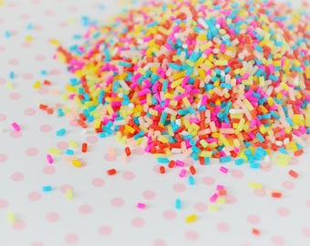Op de afbeelding: Een hoopje kleine, cilindervormige suikermuisjes in levendige tinten felroze, geel, lichtblauw, oranje en wit. De suikermuisjes vormen een dichte hoop, met losse korrels verspreid rondom de basis van de hoofdmassa.