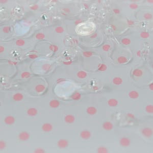 May include: A collection of clear, transparent plastic fasteners, each featuring a small circular loop connected to a slightly larger circular base. These small, lightweight components are scattered across the frame, showing a smooth, glossy finish.