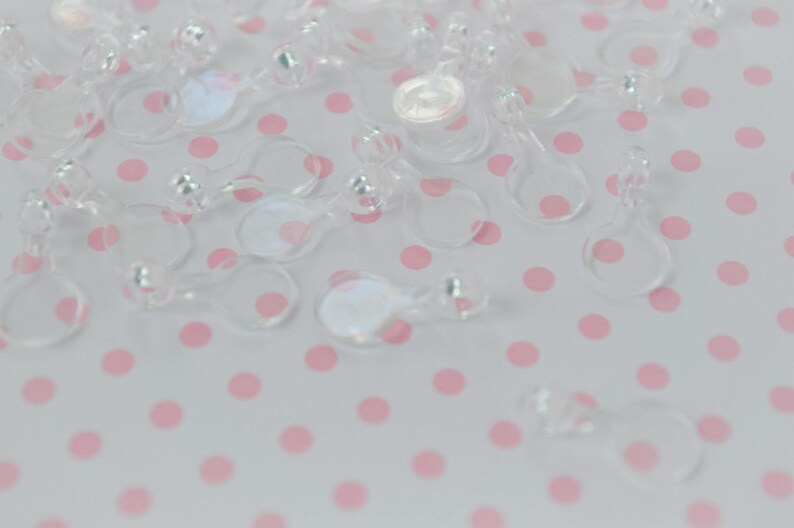 May include: A collection of clear, transparent plastic fasteners, each featuring a small circular loop connected to a slightly larger circular base. These small, lightweight components are scattered across the frame, showing a smooth, glossy finish.