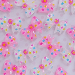 May include: A collection of translucent pink and white bow-shaped cabochons, each featuring a pattern of small blue, yellow, and pink stars. A circular pink gem with a yellow star center is positioned at the middle of every bow ornament.