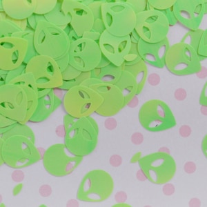 May include: Green alien face shaped confetti with a gradient from light green to dark green. The confetti is scattered on a white background with pink polka dots.
