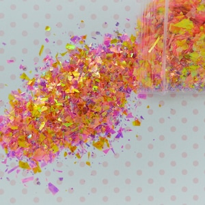 May include: A close-up of iridescent glitter in shades of pink, yellow, and orange, spilling out of a clear plastic bag onto a white background with pink polka dots.
