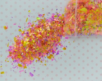 May include: A clear plastic container spilling out iridescent, multi-colored craft flakes. The thin, jagged pieces feature a reflective finish with vibrant shades of pink, orange, and yellow that shimmer and catch the light in various directions.