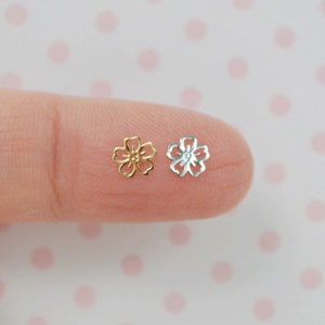 May include: Two small flower charms, one gold and one silver, are shown on a finger. The charms are delicate and intricate, with a five-petal design.