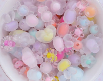 May include: A collection of assorted loose beads in various pastel colors and shapes, including flowers, hearts, bunnies, and round spheres. The beads feature a mix of translucent, frosted, and opaque finishes, creating a soft, multi-textured appearance.