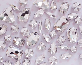 May include: A collection of clear, faceted glass rhinestones in various shapes including round, oval, pear, rectangular, and marquise cuts. These transparent, reflective gems feature sharp geometric edges and polished surfaces, scattered across the frame.