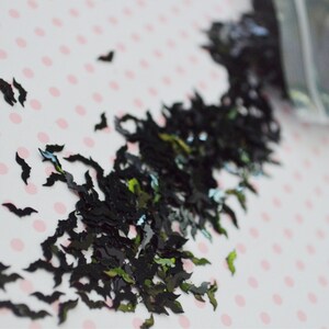 May include: Black and green glitter shaped like bats. The glitter is scattered on a white background with pink polka dots.