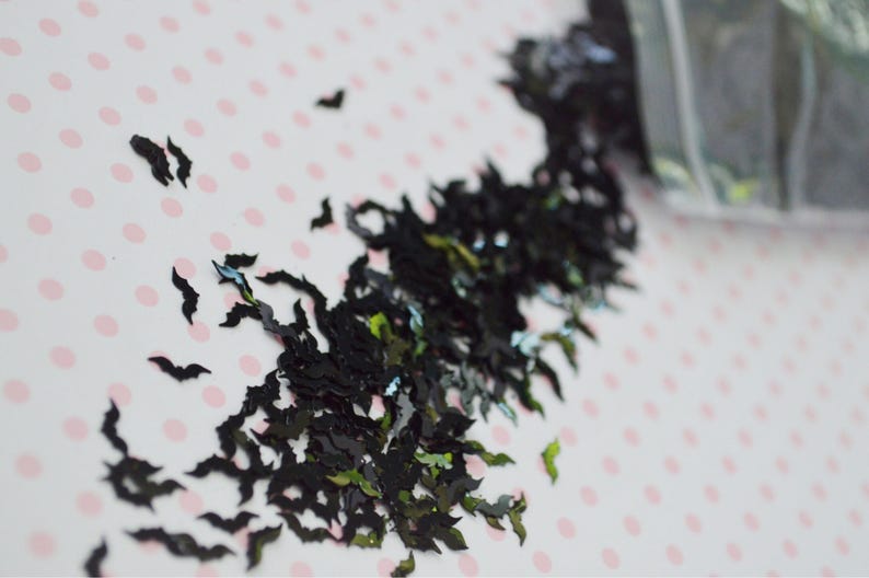 May include: Black and green glitter shaped like bats. The glitter is scattered on a white background with pink polka dots.