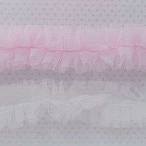 May include: Two strips of gathered tulle trim, one in a soft pastel pink and the other in a crisp white. Each strip features a central stitched line that creates a ruffled, layered effect along the edges, showcasing a delicate and sheer texture.