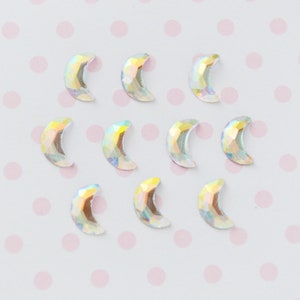 May include: A collection of nine crescent moon-shaped flatback rhinestones. Each piece features a faceted surface with an iridescent, holographic finish that reflects multiple colors including yellow, pink, and blue against a light background.