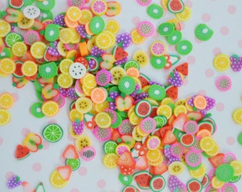 May include: A collection of small, flat, circular and triangular polymer clay slices featuring colorful fruit designs including watermelon, kiwi, orange, lemon, strawberry, and dragon fruit, scattered across a white surface with pink polka dots.