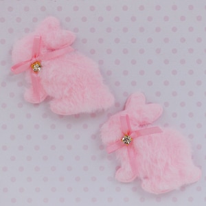 May include: Two pink fuzzy rabbit-shaped appliques, each featuring a soft plush texture. A thin pink satin ribbon is tied around the neck of each rabbit, accented with a small, clear, round rhinestone at the center of the bow knot.