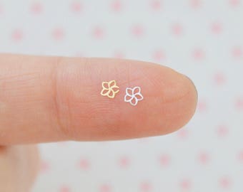 May include: Two tiny, flat, metal flower-shaped charms, one in a gold tone and one in a silver tone. Each charm features a simple, open-work outline of a five-petaled blossom, resting on a surface to show their miniature scale and delicate design.