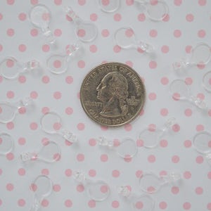 May include: A collection of small, clear plastic loop connectors scattered around a silver-colored United States quarter coin. Each connector features a circular top loop and a small, rectangular base attachment point for crafting or jewelry.