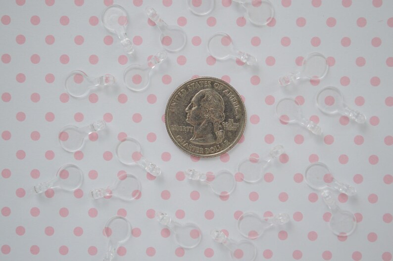 May include: A collection of small, clear plastic loop connectors scattered around a silver-colored United States quarter coin. Each connector features a circular top loop and a small, rectangular base attachment point for crafting or jewelry.