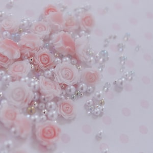 May include: A scattering of pale pink rose-shaped beads and white pearl beads on a light pink polka-dotted surface. The beads are arranged in a delicate, decorative pattern, creating a soft, romantic aesthetic.