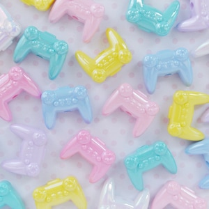 May include: A collection of pastel-colored plastic game controller charms. The charms are in various colors, including pink, blue, yellow, and white. The charms are shaped like a classic video game controller.