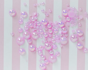 May include: A collection of round, iridescent pink beads in various sizes scattered across a surface with vertical white and light pink stripes. The beads have a glossy, reflective finish that creates a shimmering effect against the striped background.