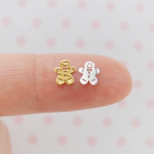 May include: Two gingerbread man charms, one gold and one silver, are shown on a finger. The charms are small and detailed, with a smiling face, arms, legs, and a bow tie.