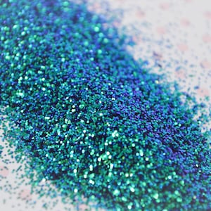 May include: A close-up of teal blue glitter with a slight iridescent sheen. The glitter is scattered on a white surface with a subtle pink polka dot pattern.