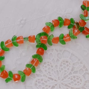 May include: A strand of glass beads shaped like orange tulips with green leaves. The beads are arranged in a repeating pattern of orange flower heads and green leaf segments, creating a continuous floral design strung together on a thin cord.