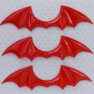 May include: Three pairs of red, shiny, bat wings. The wings are made of a flexible material and have a slightly curved shape.
