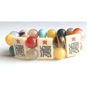 May include: A beaded bracelet featuring a mix of colorful gemstone beads and three ivory-colored Mahjong tiles with black characters. The beads are various sizes and colors, including yellow, red, blue, and brown. The bracelet is on a white background.